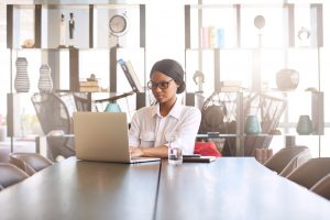 Attractive young professional well educated black woman busy typing on her notebook while seated at her dining room table at home with a large display shelf behind her in the background,.